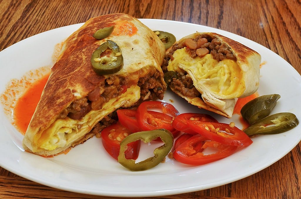 Breakfast burrito cut in half showing egg, cheese and meat, served with sliced peppers and jalapeños