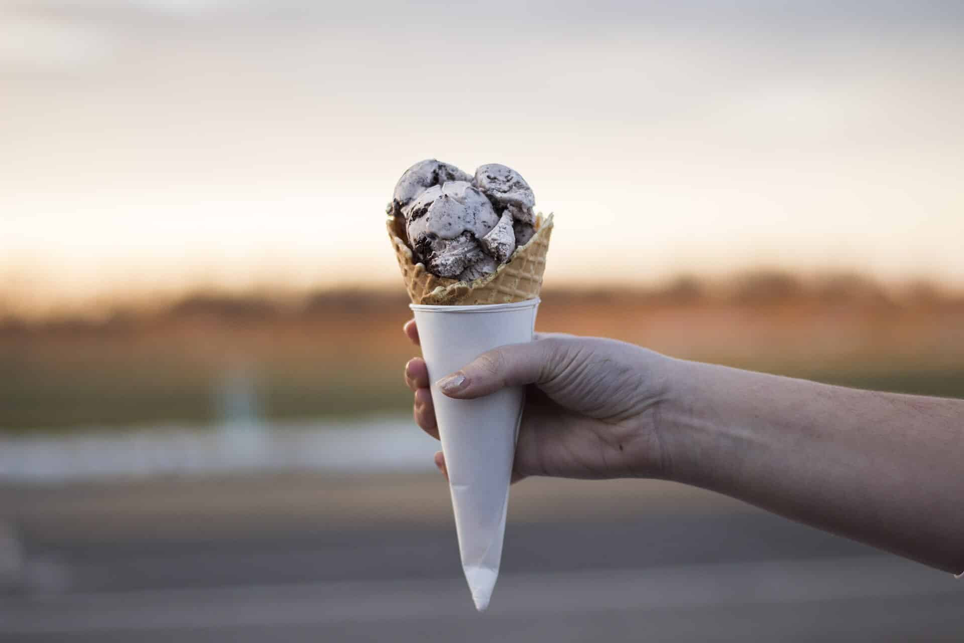 Hand holding a waffle cone piled with scoops of cookies and cream ice cream against a soft sunset