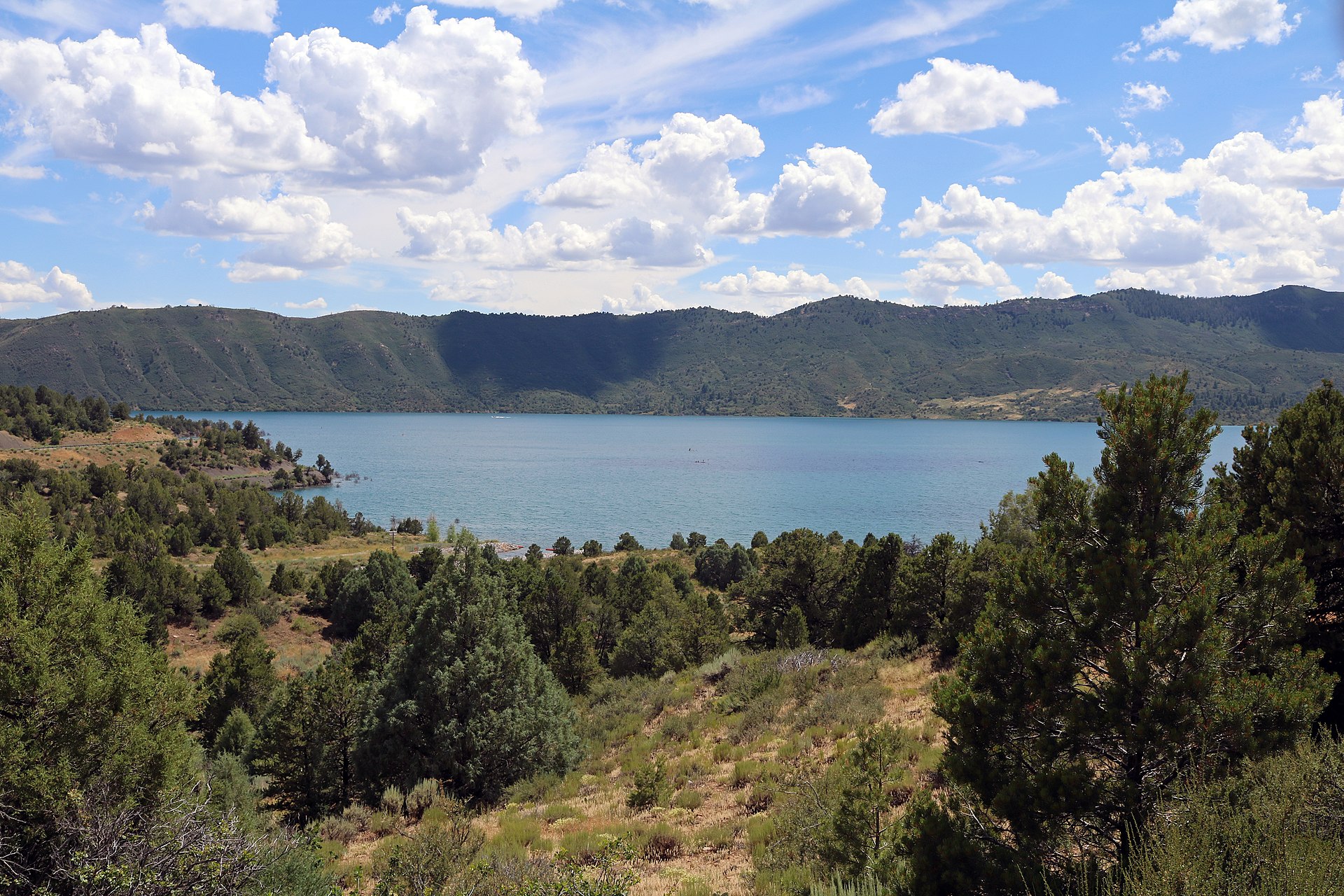 Turquoise Lake Nighthorse surrounded by forested ridges under a blue sky with white clouds, seen from a pine-dotted overlook near Durango