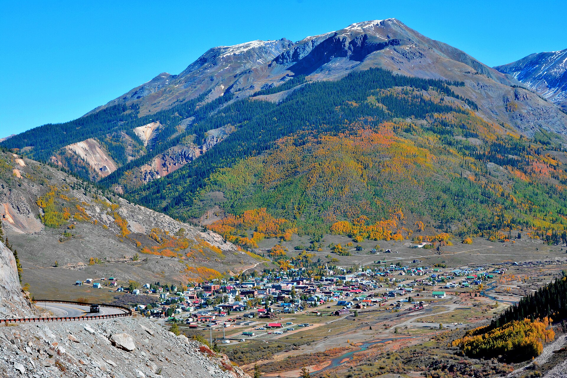The mountain town of Silverton, Colorado seen from above, surrounded by golden autumn aspens and rugged San Juan peaks under a blue sky