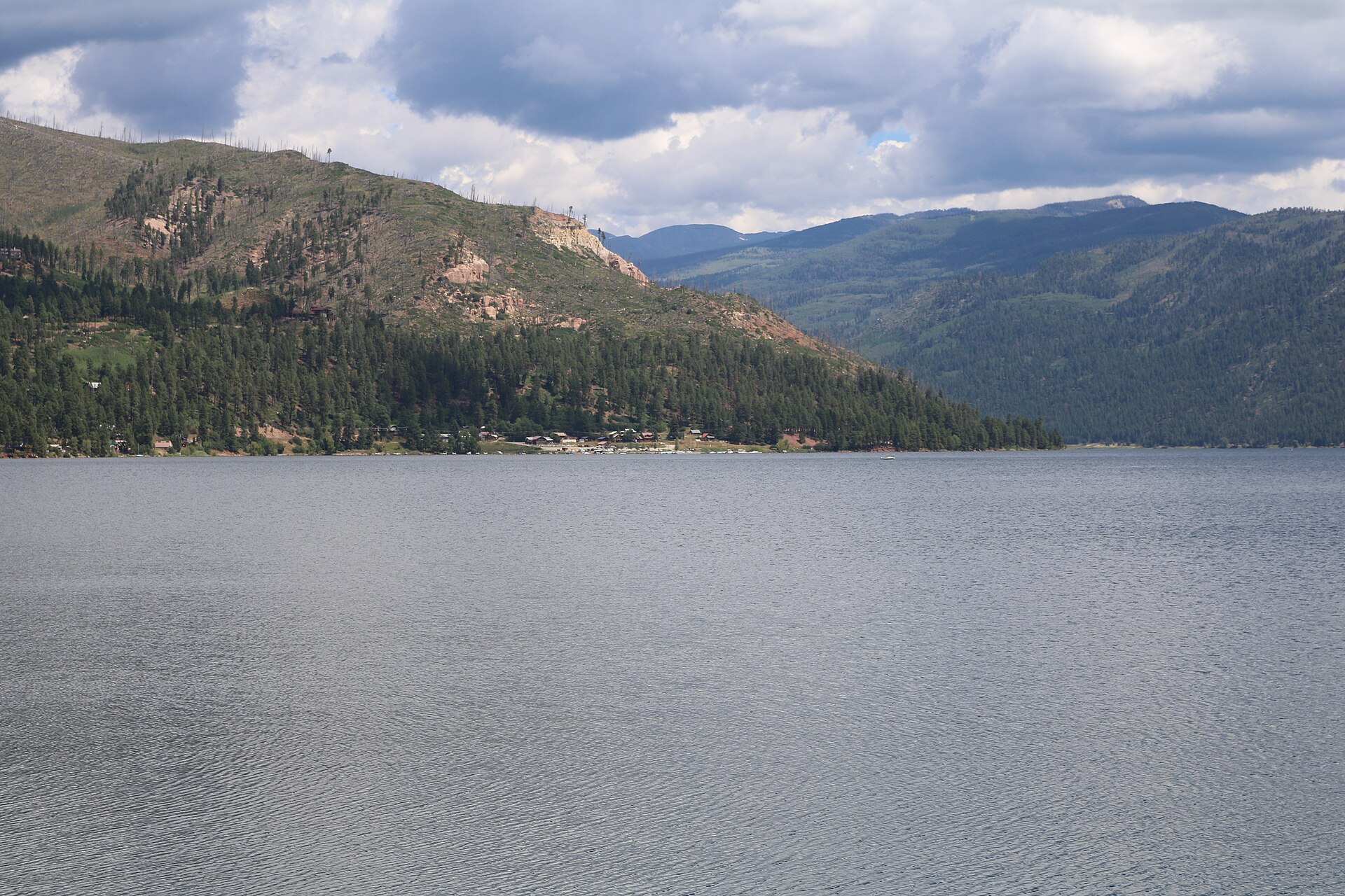 Vallecito Reservoir near Durango, Colorado, with calm water, forested hills and San Juan mountain ridges under a cloudy summer sky