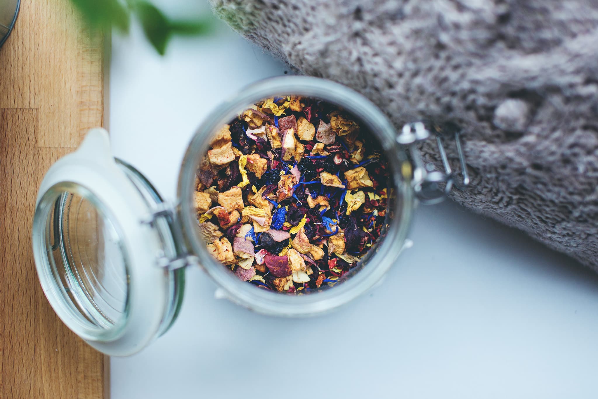 Open glass jar of loose herbal tea with dried petals and flowers, on a wooden board beside a soft grey throw